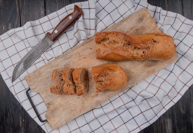 top view of half cut black baguette on cutting board with knife on plaid cloth and wooden background