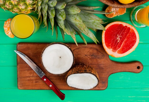 Top view of half cut coconut and knife on cutting board with grapefruit pineapple tangerine and orange juice on green background