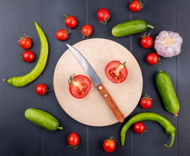 Top view of half cut tomato and knife on cutting board with pepper tomato garlic on black surface