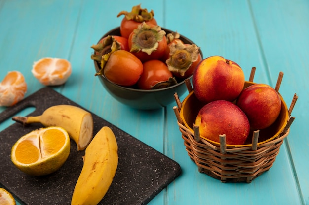 Top view of half fresh bananas on a black kitchen board with persimmons on a bowl with peaches on a bucket on a blue wooden wall