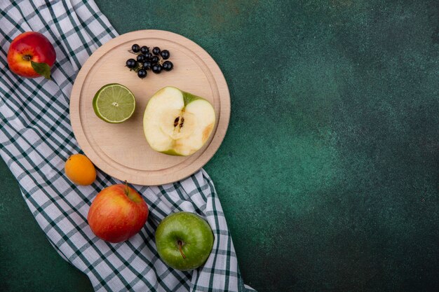 Top view of half a green apple with lime and black currants on a stand and peaches on a checkered towel on a green surface