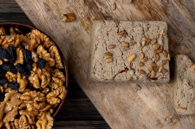Top view of halva with sunflower seeds and walnuts in a bowl on a wooden board