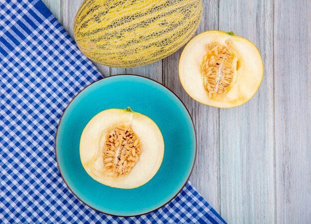 Top view of halved melons on a blue plate on blue checked tablecloth on grey wooden surface