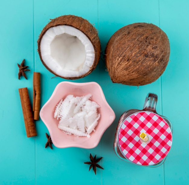 Top view of halved and whole coconuts with pulps of coconut in a pink bowl with cinnamon and anise juice in glass jar on blue surface