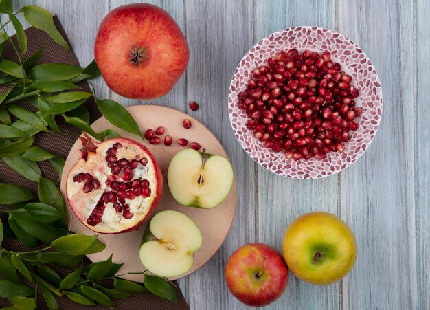 Top view of halves of a green apple on a stand with peeled pomegranate in a plate with leaf branches on a gray surface