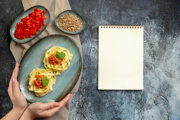 Вид сверху руки, держащей синюю тарелку с вкусной пастой, подаваемой с помидорами и мясом на ужин на коричневом полотенце, его ингредиенты рядом со спиральной записной книжкой