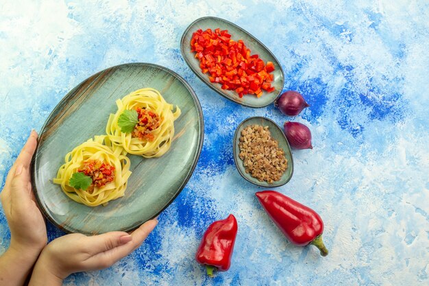 青いテーブルにおいしいパスタと必要な野菜の肉と青いプレートを持っている手の上面図