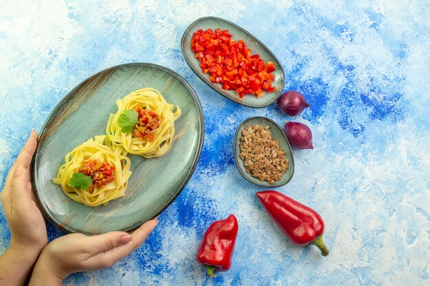 파란색 테이블에 맛있는 파스타와 필요한 야채 고기가 있는 파란색 접시를 들고 있는 손의 상단