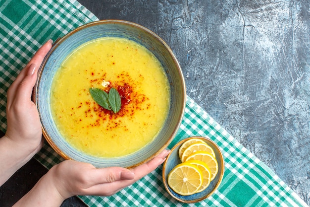 Top view of hand holding a blue pot with tasty soup served with mint and chopped lemon on half folded green stripped towel on blue background