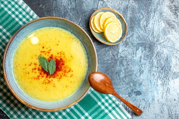 Top view of hand holding a blue pot with tasty soup served with mint and pepper next to chopped lemon wooden spoon on blue background