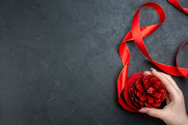 Top view of hand holding a conifer cone with red ribbon on dark background