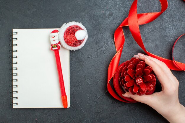 Top view of hand holding a conifer cone with red ribbon and notebook with red ribbon on dark background