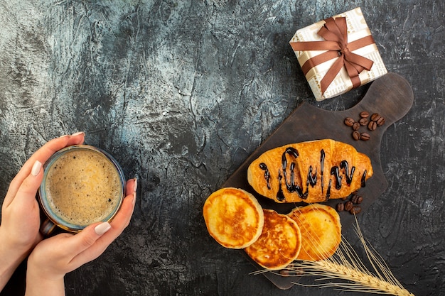 Top view of hand holding a cup of coffee and tasty breakfast with pancakes croisasant and gift box on dark table