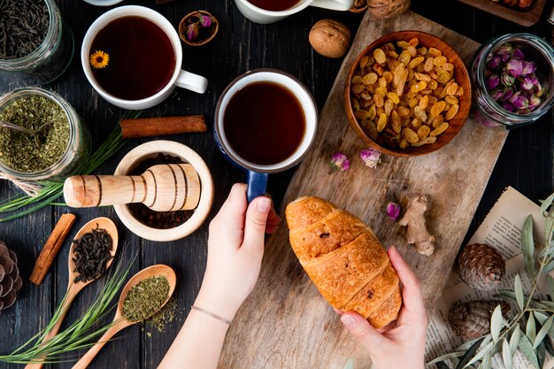 Top view of hand holding a cup of tea and croissant over the wooden board with dried raisins in a bowl and various spices and herbs on wood