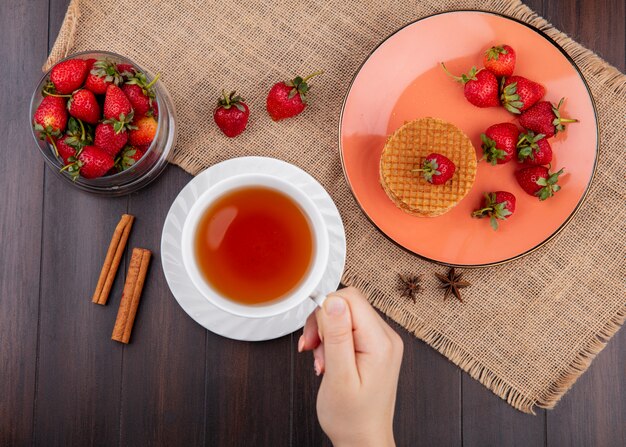 木の表面にシナモンと荒布を着たお茶とワッフルビスケットのプレートとイチゴのボウルを持っている手の平面図