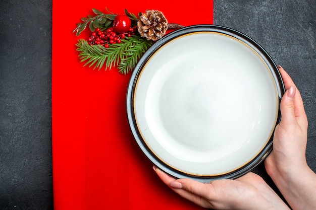 Top view of hand holding empty plates and fir branches with decoration accessory conifer cone on a red napkin on a dark background