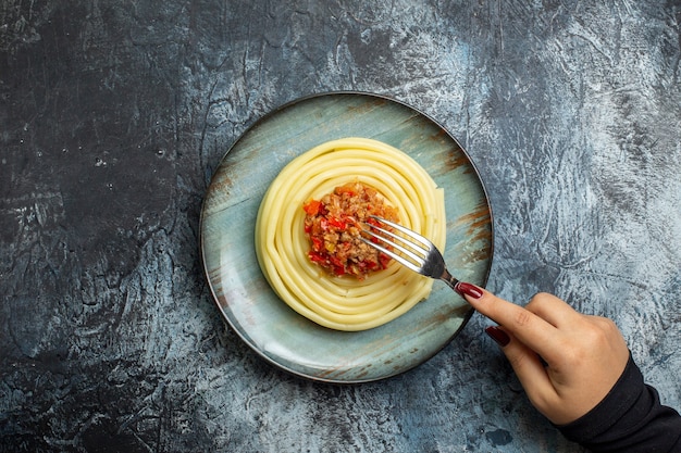 Top view of hand holding fork on delicious pasta meal on a blue plate served with tomato and meat for dinner