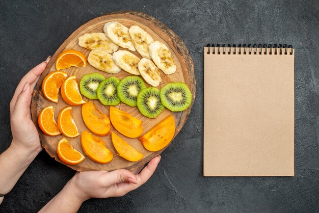 Top view of hand holding a natural organic fresh fruit set on cutting board and closed notebook on dark surface