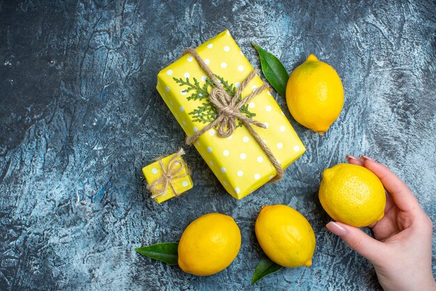 Top view of hand holding one of fresh lemons with leaves and yellow gift boxes on dark background