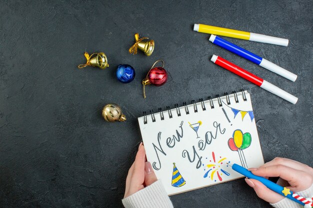 Вид сверху руки, держащей ручку на спиральной записной книжке с новогодними украшениями для письма и рисунков на черном фоне