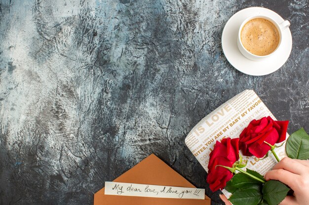 Top view of hand holding red roses on beautiful gift box and cup of coffee envelope with love letter on icy dark background