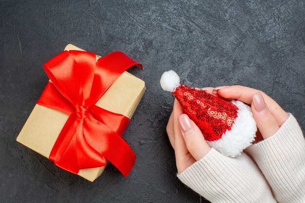 Top view of hand holding santa claus hat and beautiful gift with bow-shaped ribbon on dark background