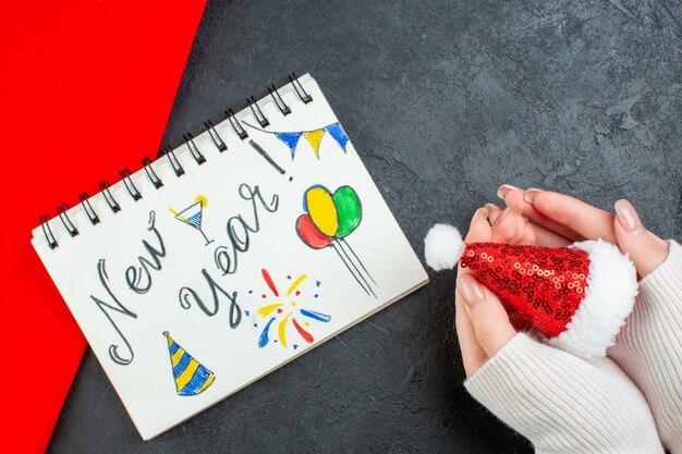 Вид сверху руки, держащей шляпу санта-клауса и блокнот с красным полотенцем с новогодним письмом и рисунками на темном фоне