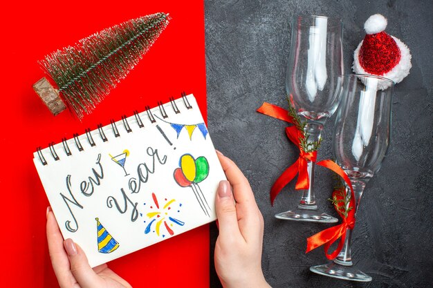 Вид сверху руки, держащей спиральный блокнот с новогодним рисунком и стеклянными кубками рождественской елки на темном и красном фоне