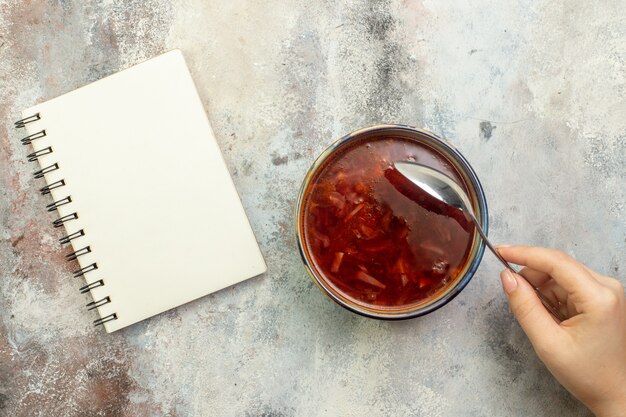 カラフルな背景においしいボルシチのスープとスパイラルノートと青い鍋にスプーンを持っている手の上面図