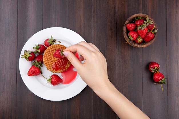 Top view of hand holding strawberry with waffle biscuits in plate and bowl of strawberry on wooden surface