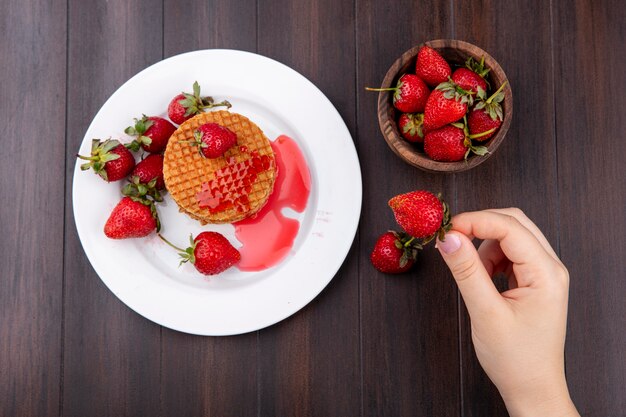 Top view of hand holding strawberry with waffle biscuits in plate and bowl of strawberry on wooden surface