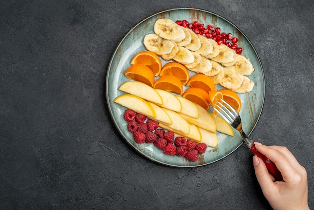 Top view of hand taking apple slices from with fork collection of chopped fresh fruits on a blue plate on black table
