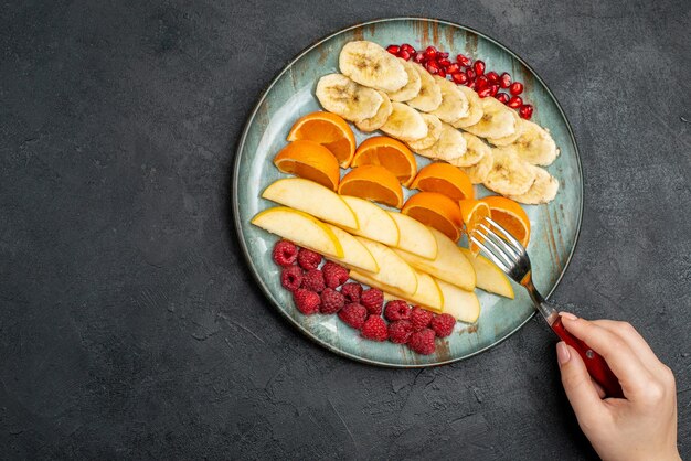 Top view of hand taking apple slices from with fork collection of chopped fresh fruits on a blue plate on black table