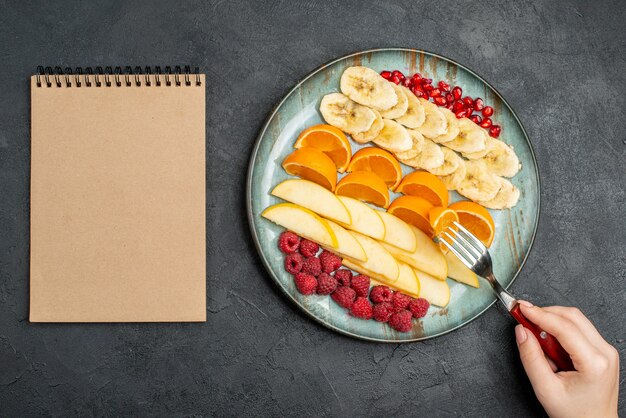 Top view of hand taking apple slices from with fork collection of chopped fresh fruits on a blue plate and spiral notebook on black table