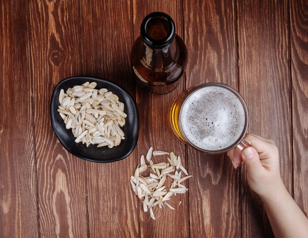 Top view of a hand with a mug of beer and a bottle of beer with salty snack sunflower seeds in a bowl on rustic wood