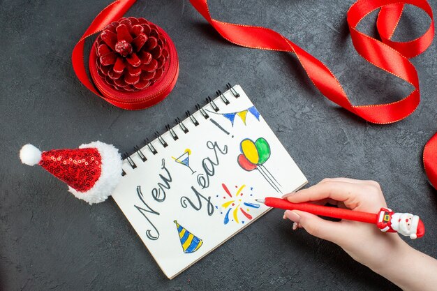 赤いリボンと暗い背景に新年の書き込みとサンタクロースの帽子とノートブックで針葉樹の円錐形を書いている手書きの上面図