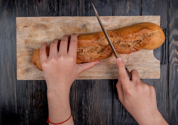 top view of hands cutting black baguette with knife on cutting board on wooden background