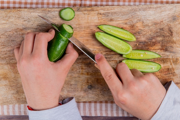 Top view of hands cutting cucumber with knife on cutting board on plaid cloth