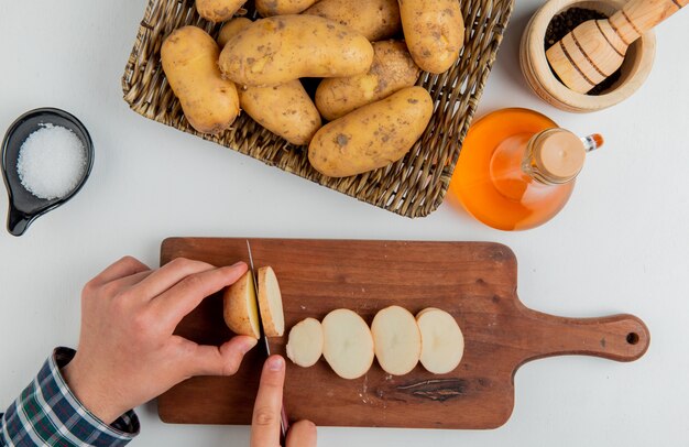 Вид сверху руки режут картофель с ножом на разделочную доску и другие в тарелку масло соль черный перец на белой поверхности
