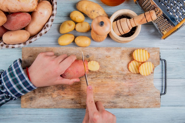 Top view of hands cutting potato with knife on cutting board with other ones in basket with black pepper seeds grater and other potatoes on wooden surface