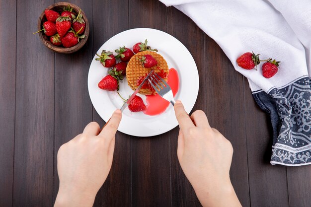 Top view of hands cutting waffle biscuit with fork and knife in plate and strawberries on cloth and in bowl on wooden surface