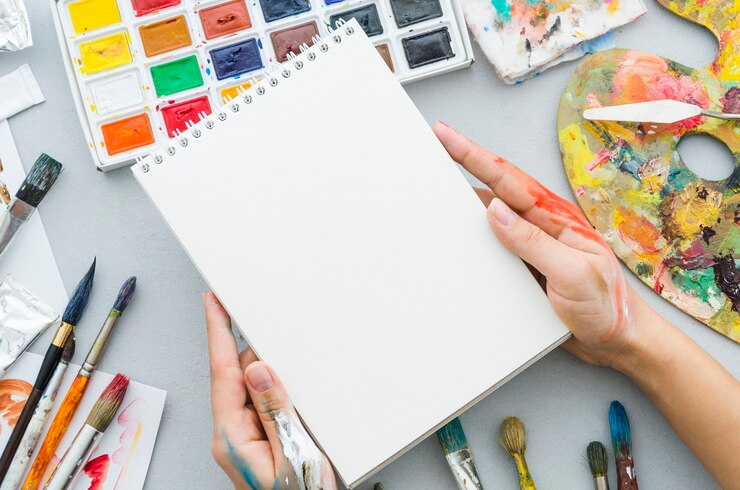 Free Photo top view hands holding notebook surrounded by painting elements