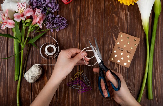 Top view of hands with scissors cutting a rope postcard paper clips and a bouquet of pink alstroemeria flowers with lilac on wooden background