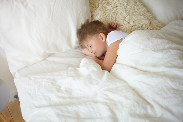 学校でのレッスンの後に昼寝をしているヨーロッパの外観のハンサムな愛らしい男子生徒の上面図。白いシーツの上にベッドで安らかに眠っている白いTシャツの甘い魅力的な男の子、眠っている笑顔