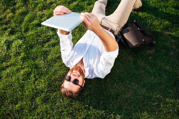Top view of happy bearded man in sunglasses and business clothes lying on grass outdoors and making selfie on tablet computer