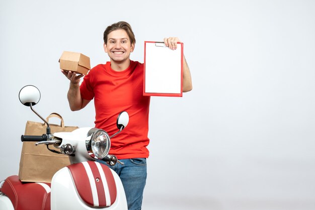 Top view of happy delivery guy in red uniform standing near scooter holding order and document on white background