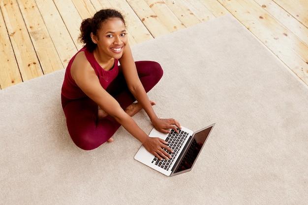 Top view of happy energetic young dark skinned woman with beaming smile sitting on carpet with generic portable computer, checking email after indoor training. Sports and active lifestyle concept