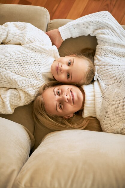 Top view of happy family relaxing at home. Vertical shot of beautiful young mother and her cute daughter lying head to head next to each other on sofa, smiling happily, wearing sweaters