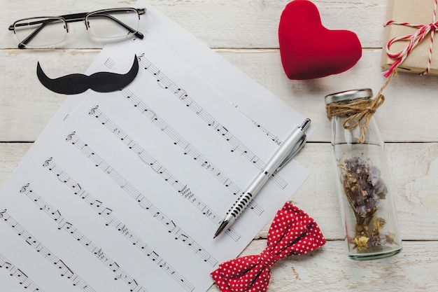 Free photo top view happy father day with music concept.music note paper on rustic wooden background.accessories with red heart,gift,mustache,vintage bow tie,dried flower in the bottle and present.