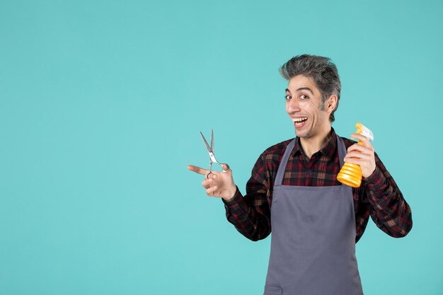 Top view of happy male barber wearing gray apron and holding scissors spray bottle on soft blue color background